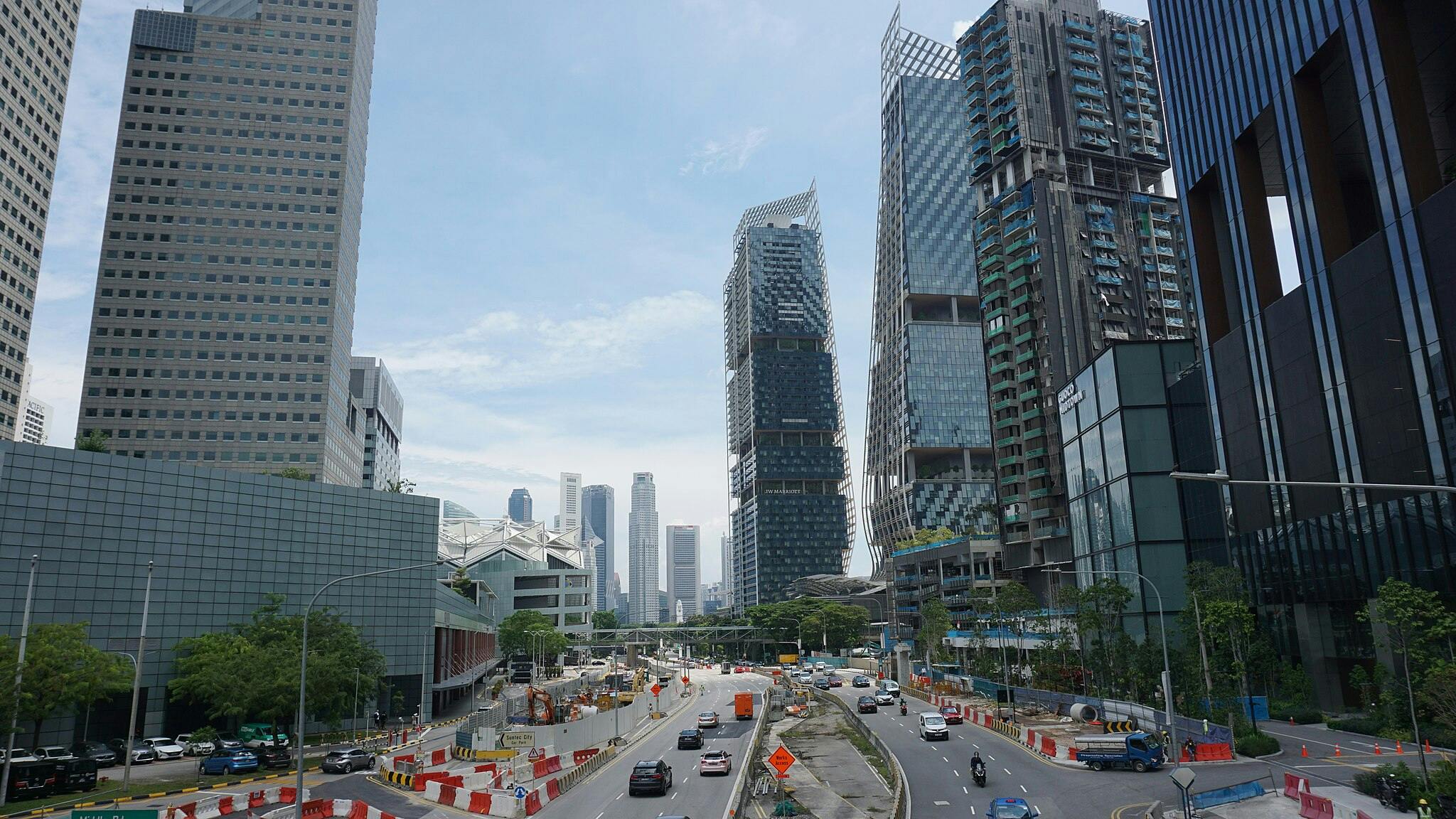 Městská ulice s auty, obklopená vysokými moderními budovami, stavebními bariérami a jasně modrou oblohou.