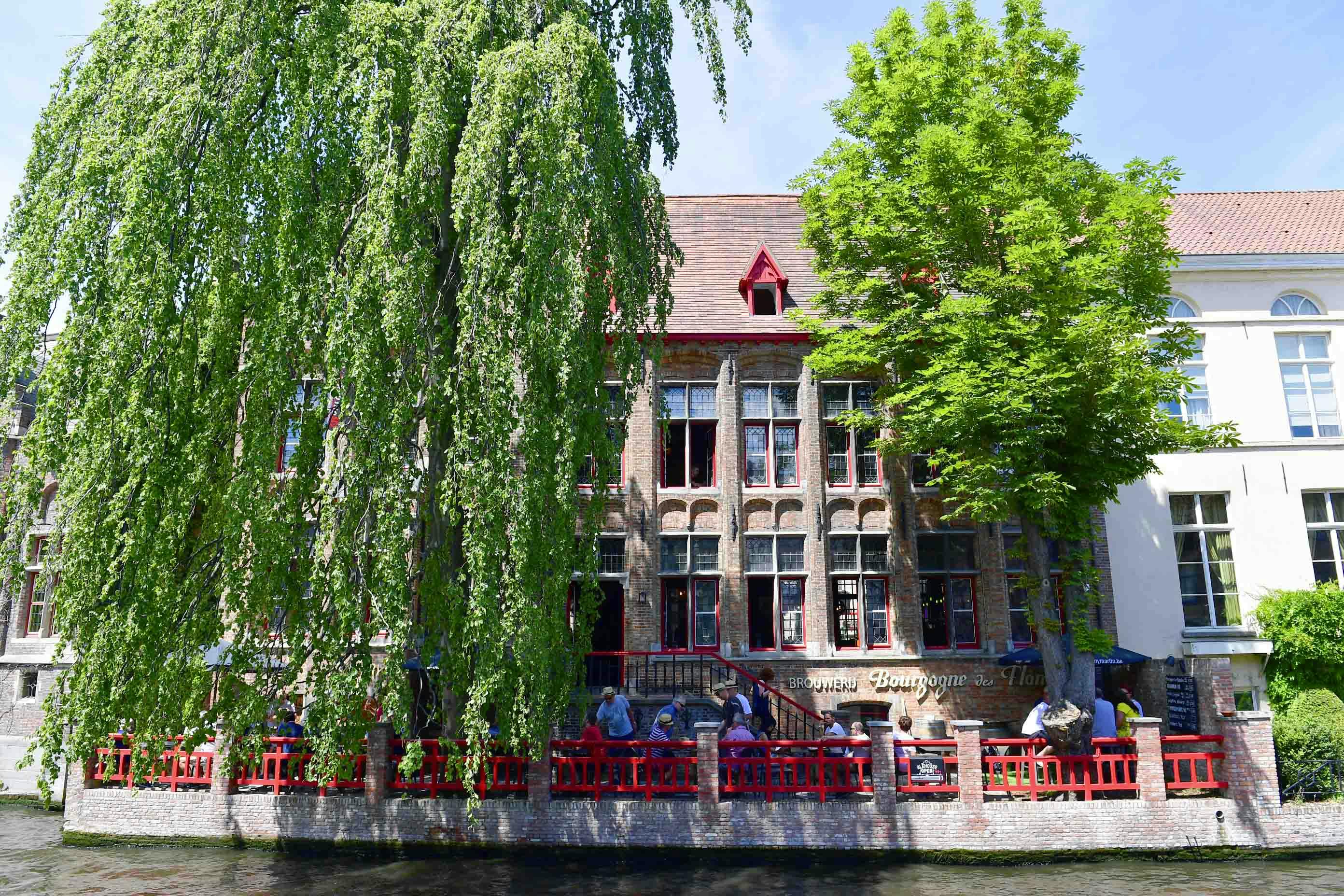 Ludzie siedzą w kawiarni na świeżym powietrzu z czerwonym ogrodzeniem, przy kanale, naprzeciwko zabytkowego budynku z wysokimi oknami i bujnymi drzewami.