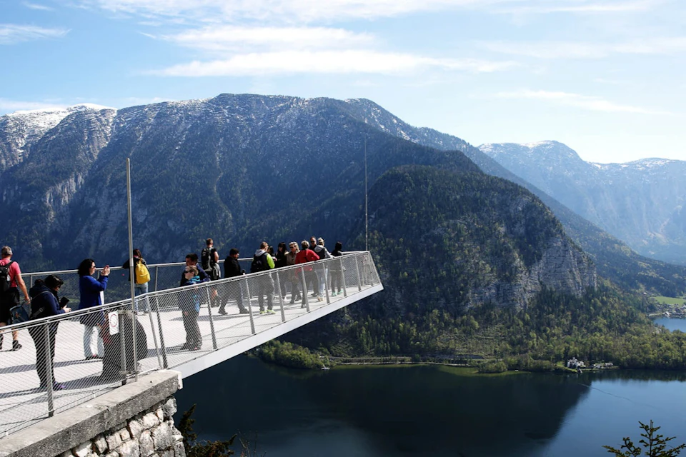 People stand on a metal viewing platform extending over a mountainside, with a lake and forest visible below.