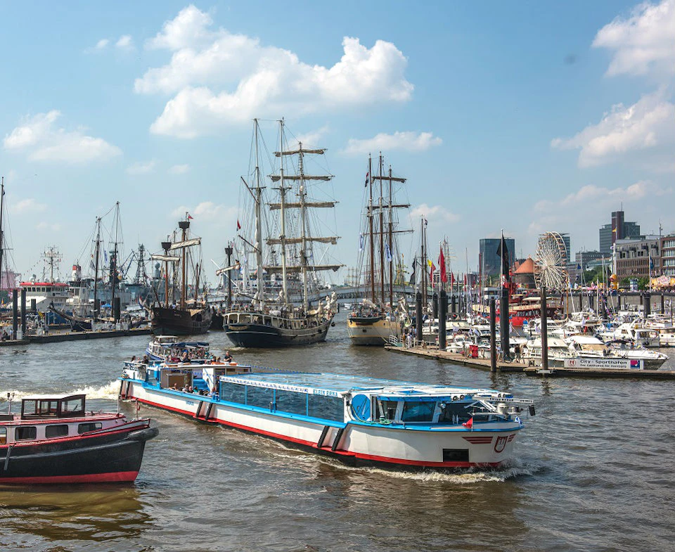 A busy harbor scene with various boats, including tall ships, a long blue and white vessel, and numerous docked sailboats.