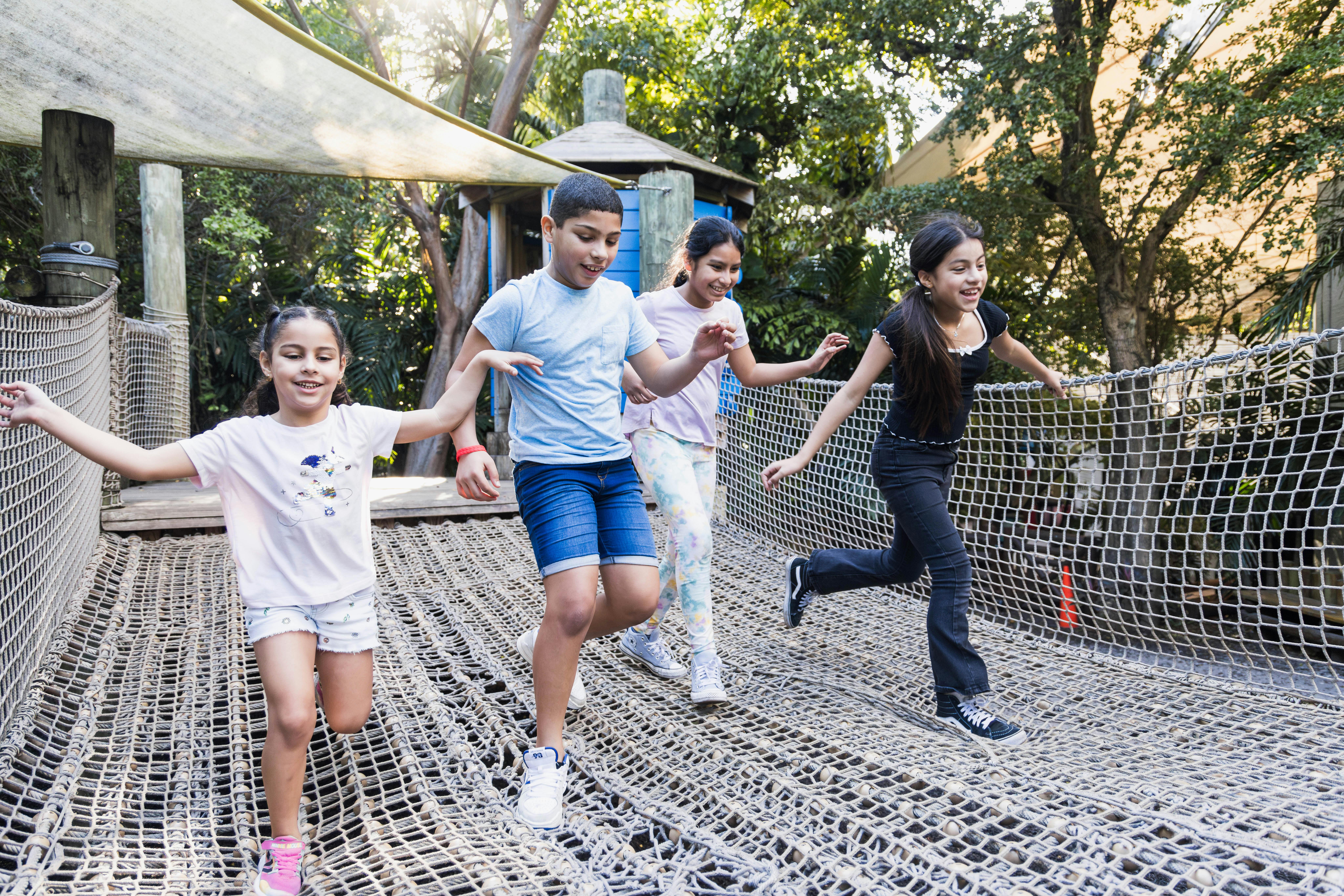 木々や木造の建物に囲まれた公園の高架橋の上で、4人の子供たちが笑顔とバランスを取りながら遊んでいます。