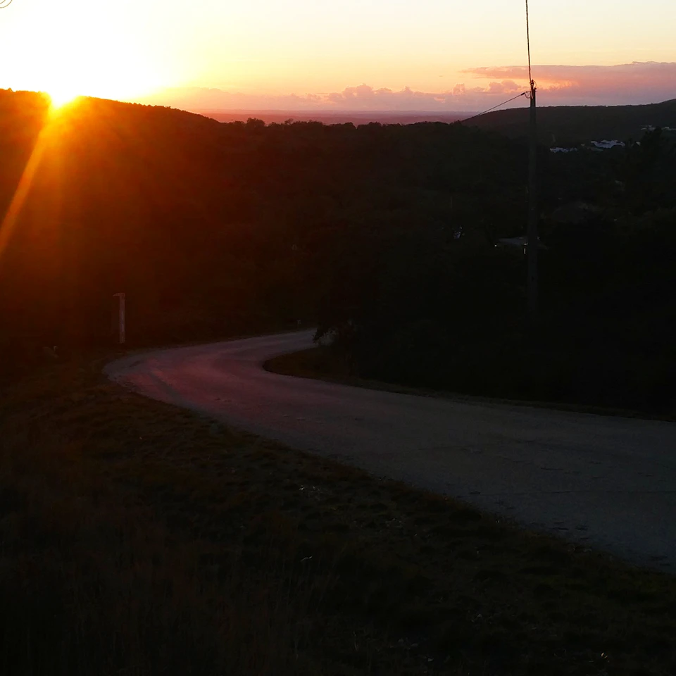 A winding road curves through a dimly lit landscape, illuminated by a setting sun peeking over distant hills.