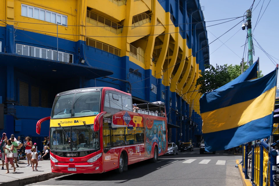 A red double-decker tourist bus parked near a bright blue and yellow building on a sunny day.