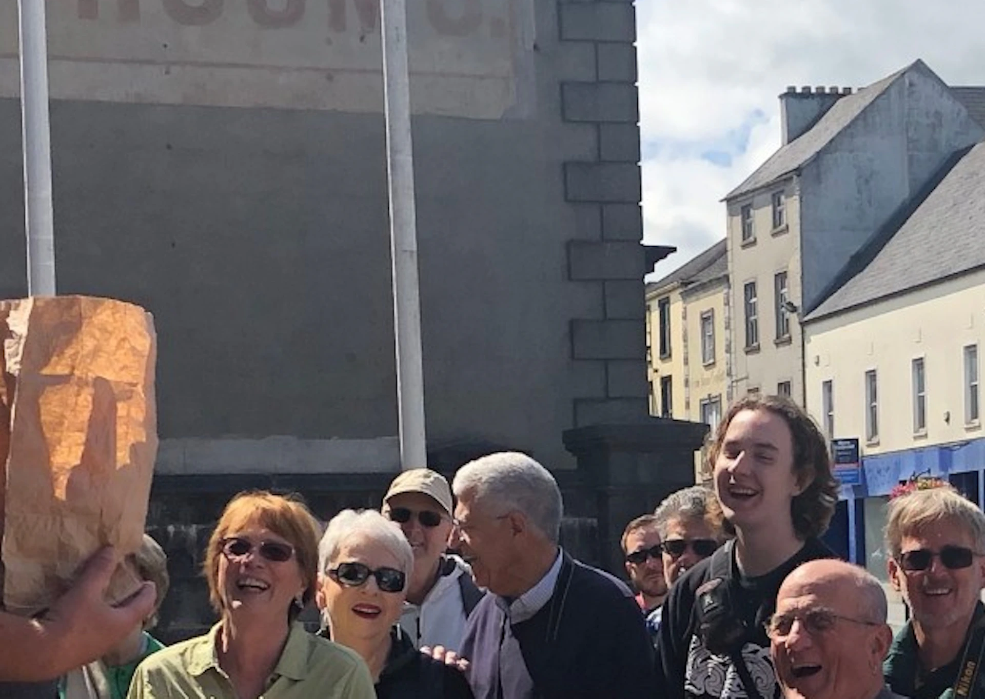 A man holding a paper bag speaks to a crowd of smiling onlookers on a sunny day in a town square.