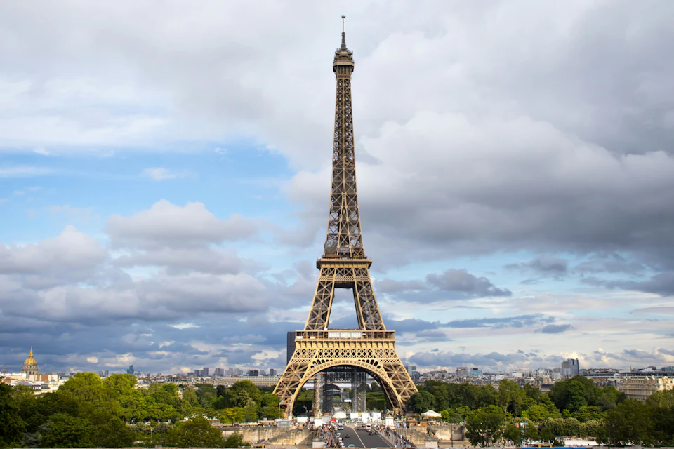 The Eiffel Tower in Paris under a cloudy sky, with trees and cityscape in the background.