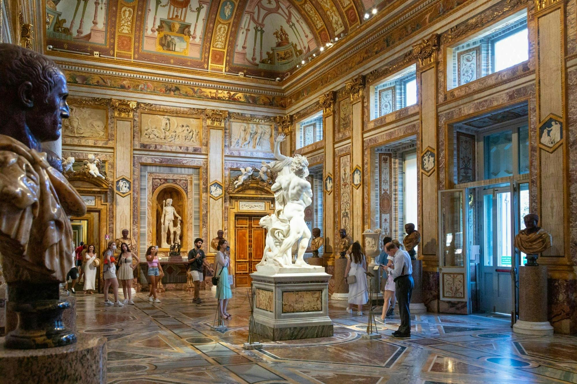 People observing marble statues in an ornately decorated museum hall with high ceilings, large windows, and intricate artwork.