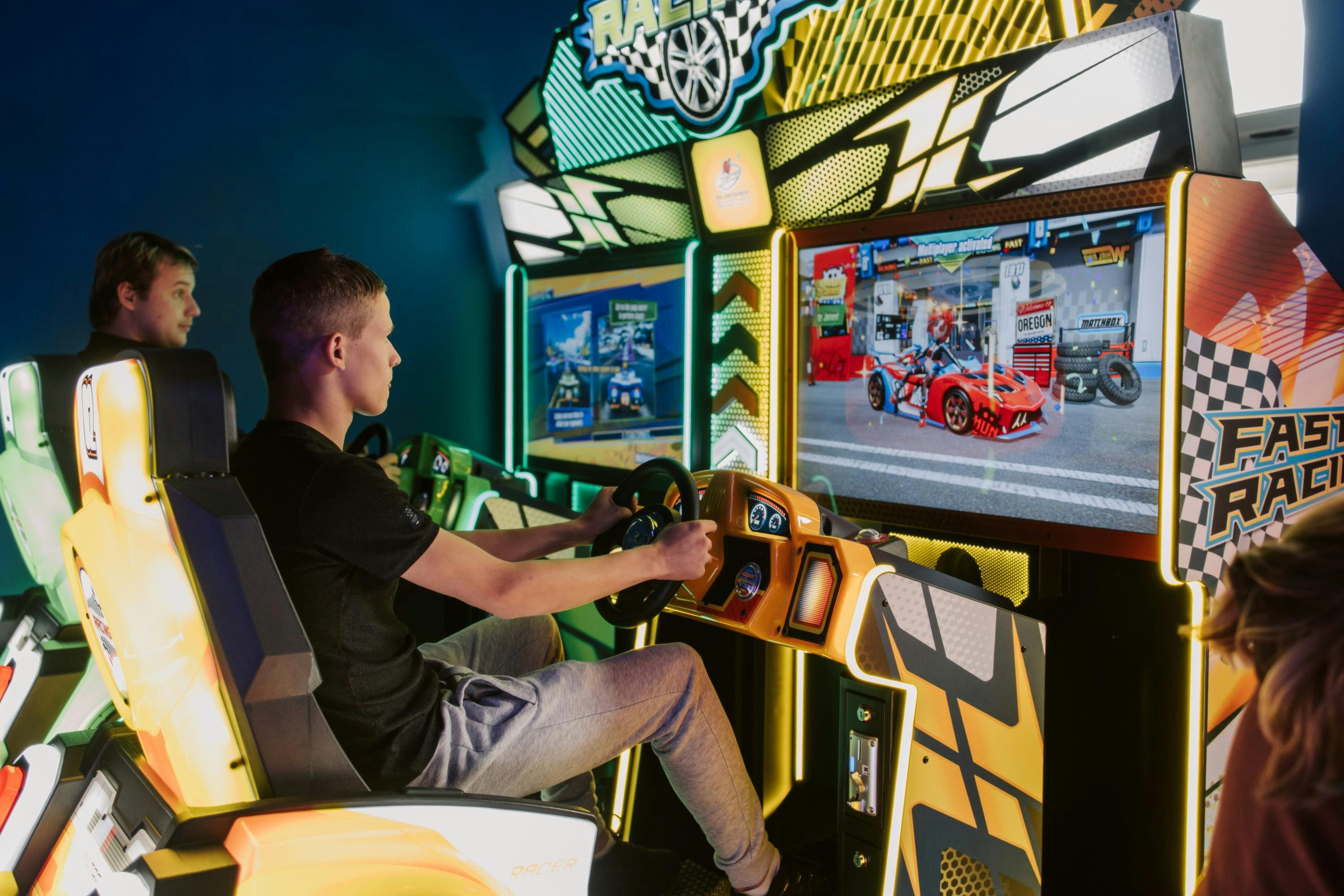 Two people playing an arcade racing game, seated in simulation cars with a bright game screen in front of them.