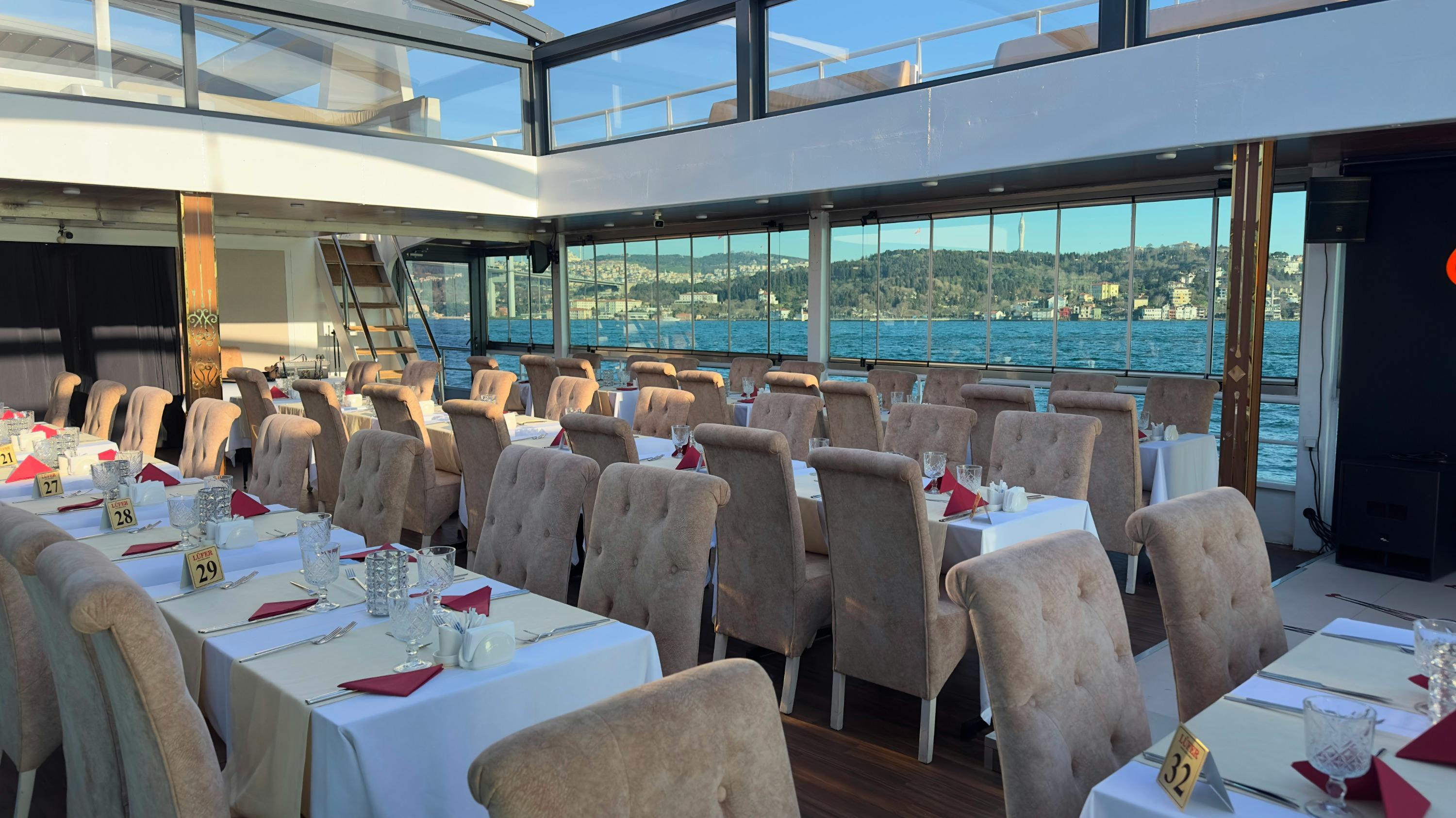 A dining area with neatly arranged tables and chairs, large windows showing a water view and distant landscape.