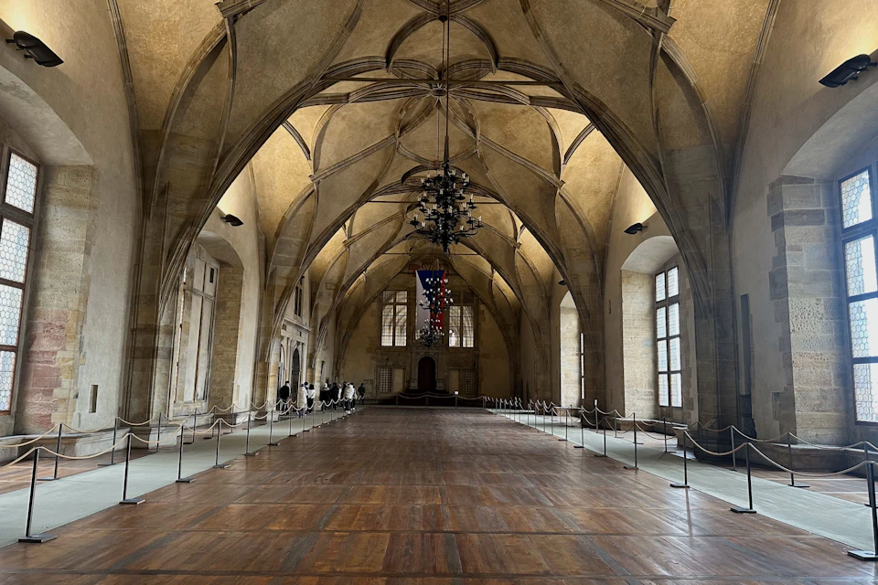 A grand hall with a wooden floor, vaulted ceiling, chandeliers, and large arched windows, with a banner hanging centrally.
