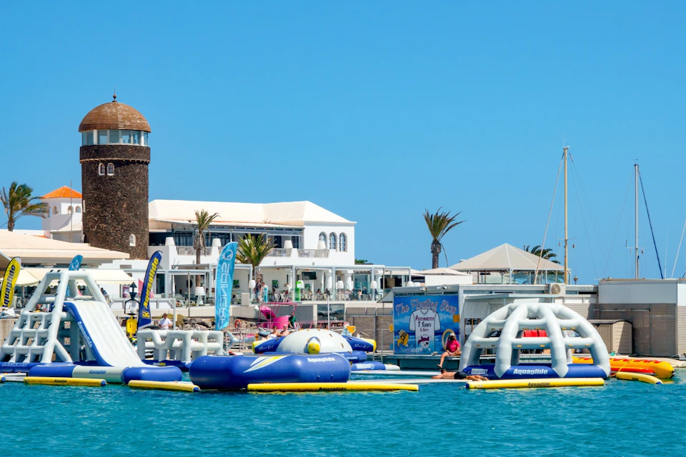 Floating water park with inflatable structures in front of a coastal building and palm trees under a bright blue sky.