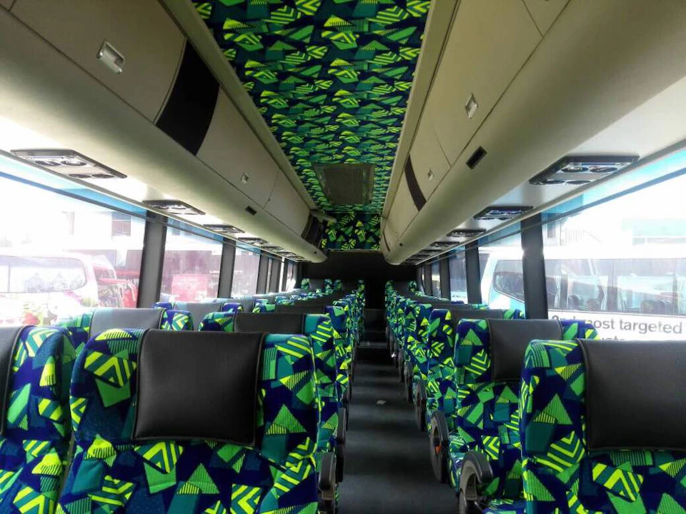 Interior of a bus with patterned green and blue seats and overhead luggage compartments.