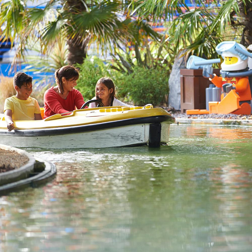 三个人乘坐黄色船只，四周环绕着棕榈树，附近有一个蓝色和橙色的机器人雕像。