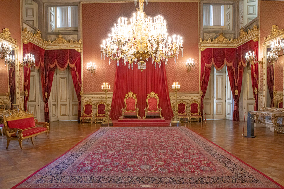 Ornate room with red curtains and grand chandelier, featuring two elaborately decorated gold chairs on a raised platform.
