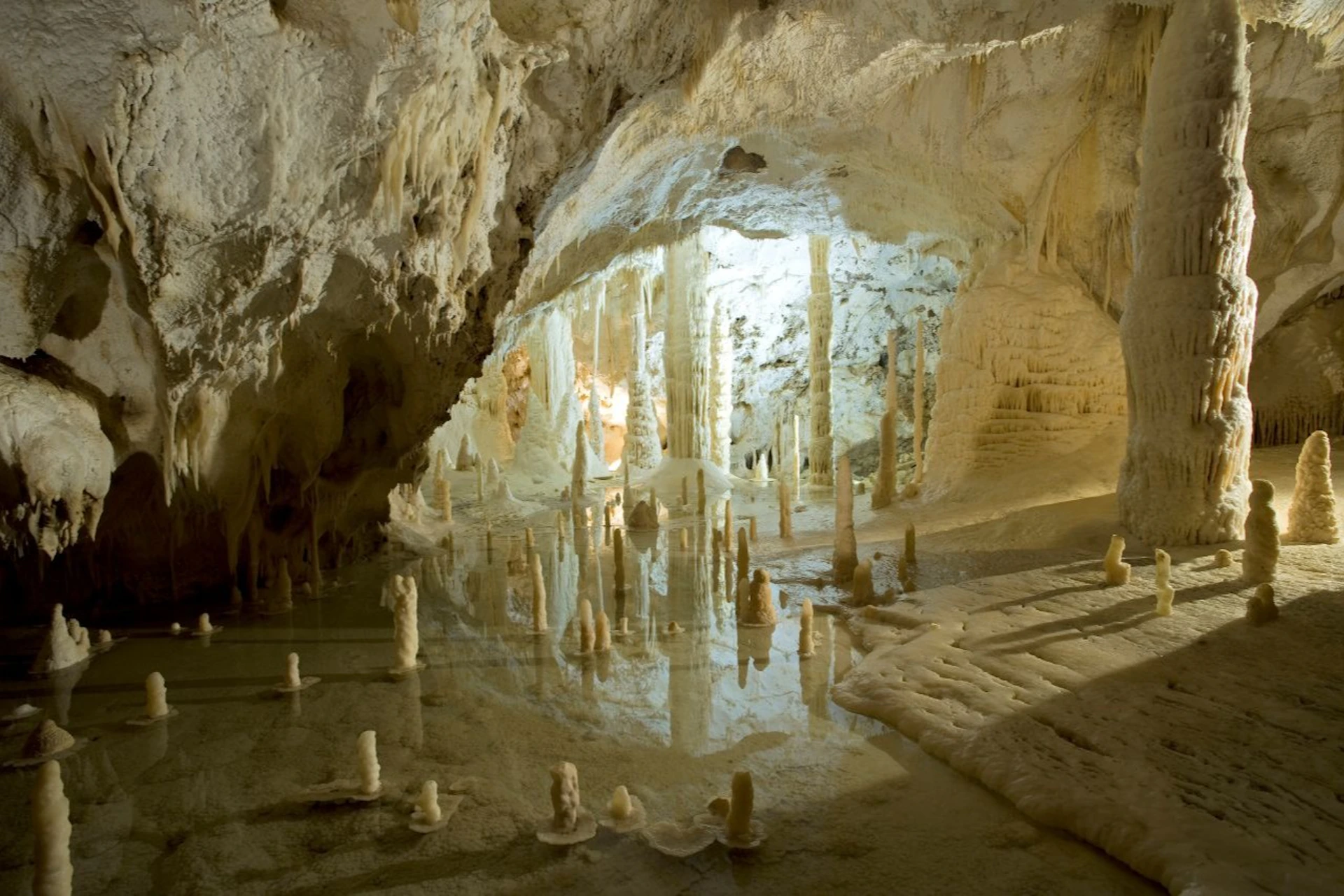 A limestone cave interior with stalactites and stalagmites reflecting in a pool of water, illuminated by artificial lighting.