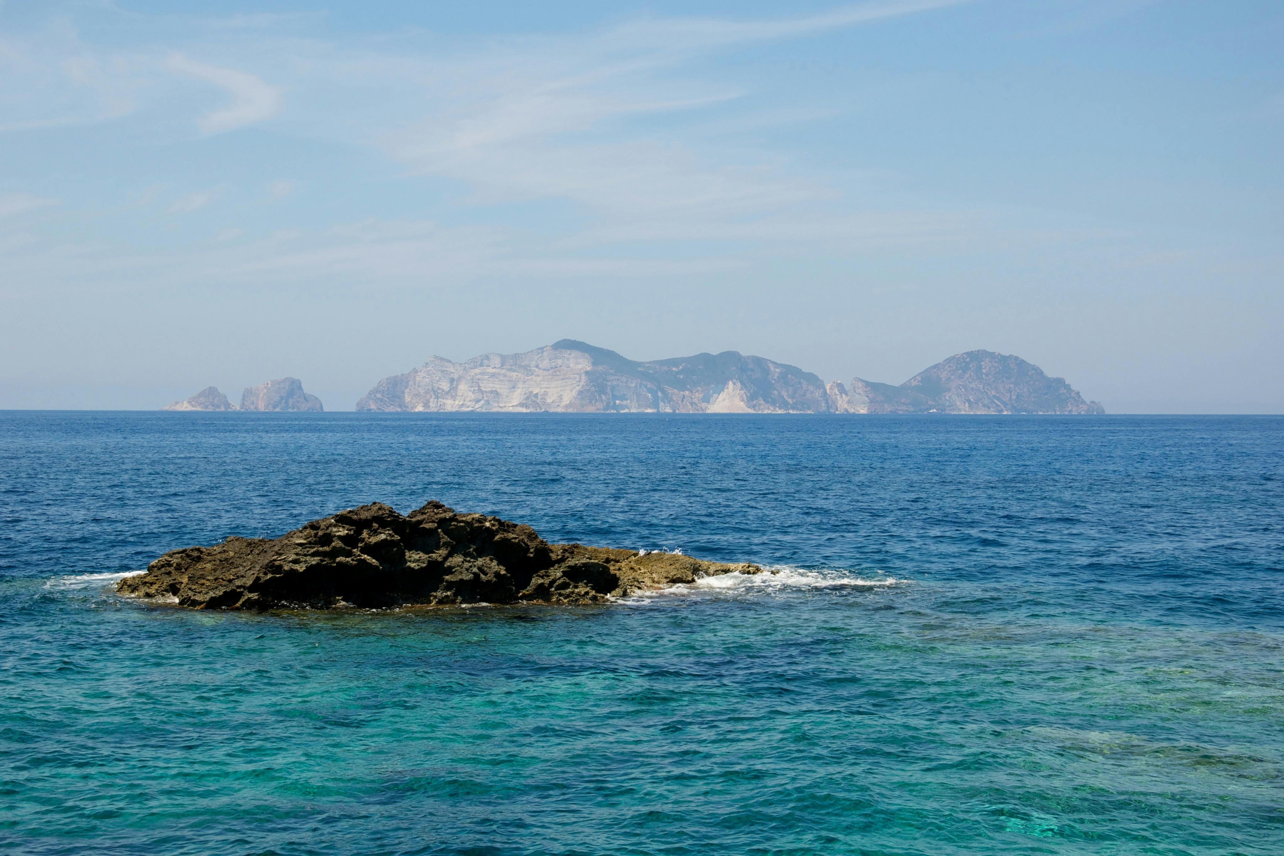 Rotsen in een helderblauwe zee met ruige eilanden in de verte onder een lichtbewolkte hemel.