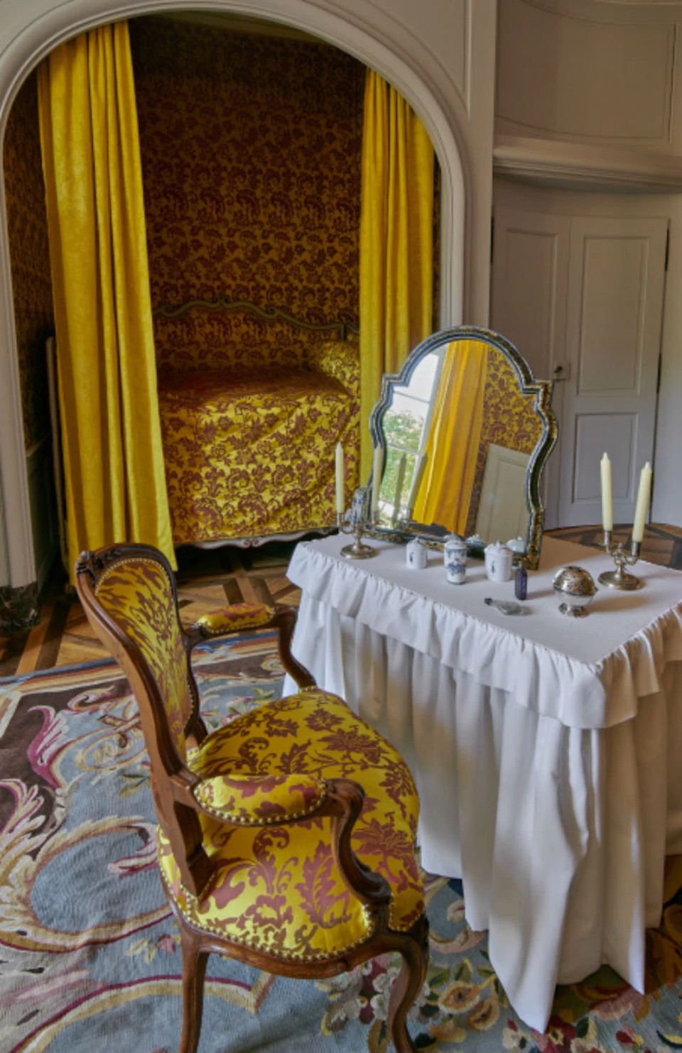 Ornate bedroom with yellow and red patterned wallpaper, canopy bed, matching chair, and a vanity table with a mirror and candles.