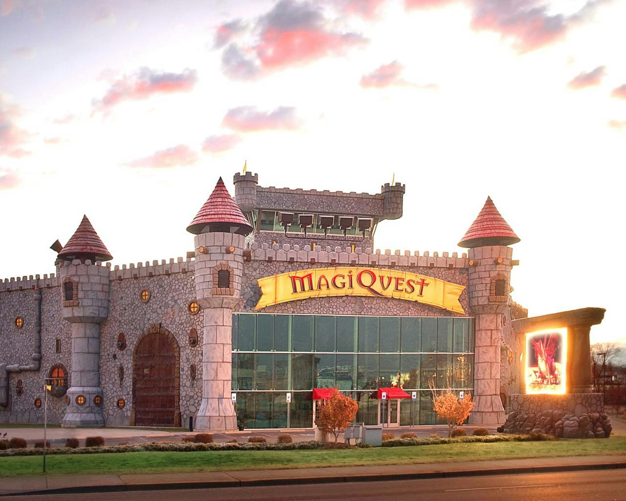 Stone castle-themed building with red-roofed towers and a sign reading "MagiQuest" above the glass entrance.