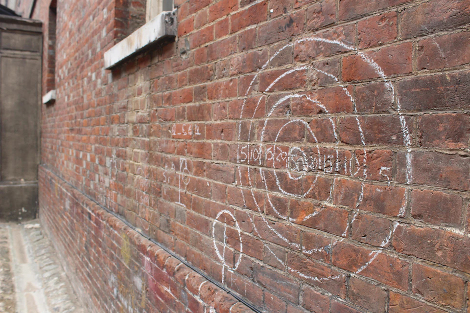 Brick wall with chalk-drawn concentric circles, numbers, and a small figure near a window ledge.