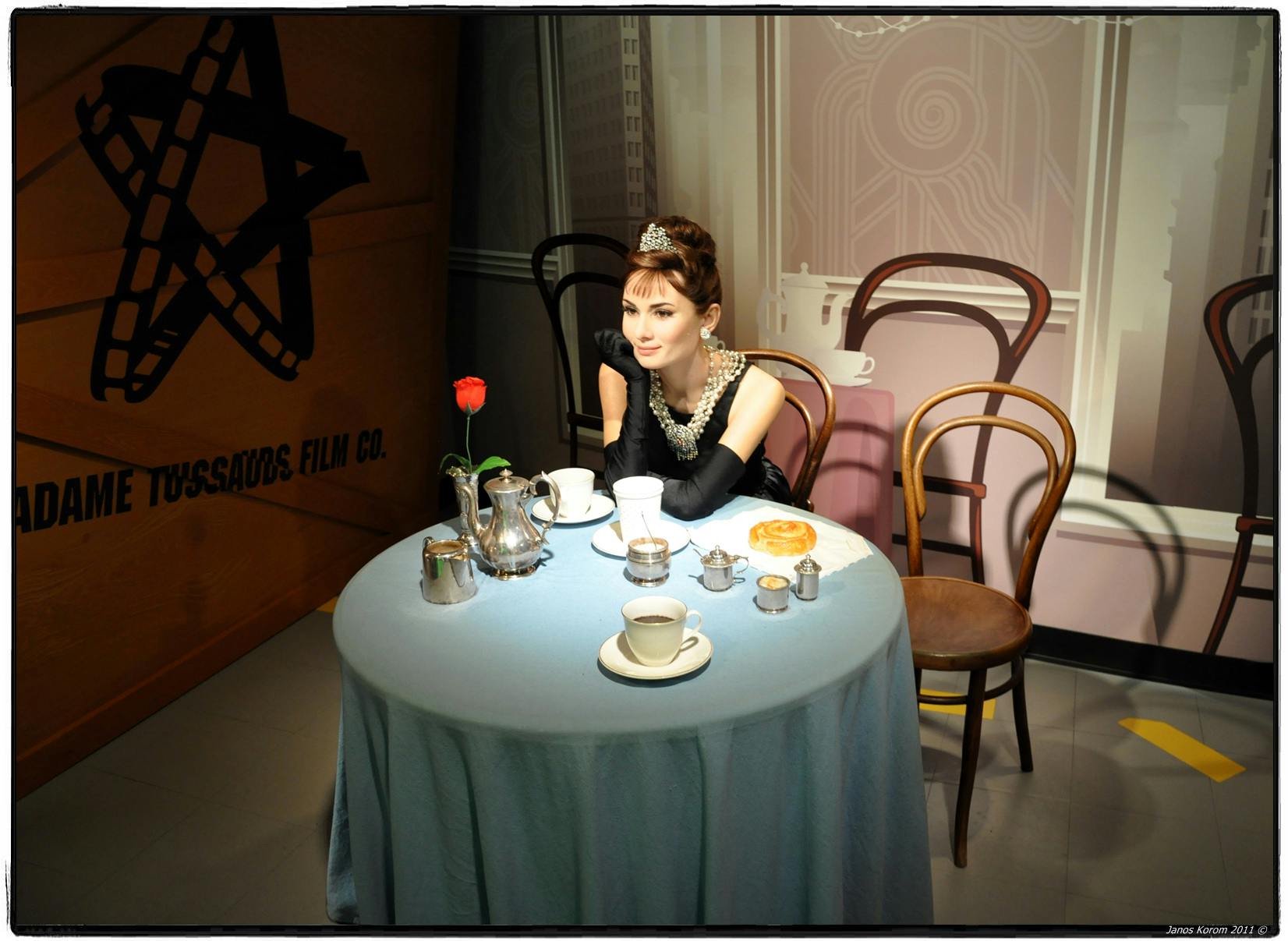 Una figura de cera d'una dona amb un vestit elegant s'asseu a una taula amb un joc de te, una pastisseria i una sola rosa vermella.