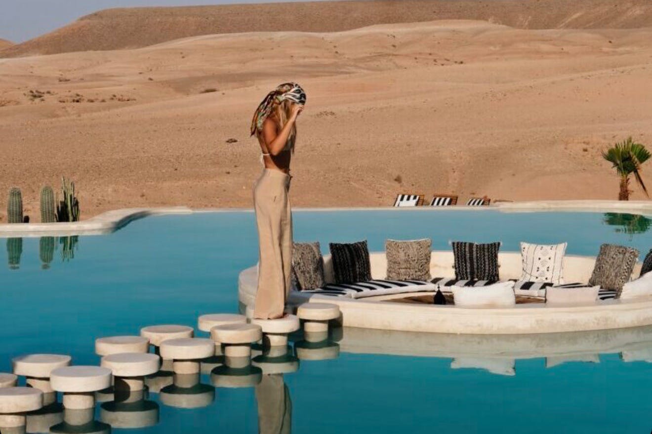 A person stands on circular stepping stones over a pool, near a round lounge area with cushions, set against a desert backdrop.