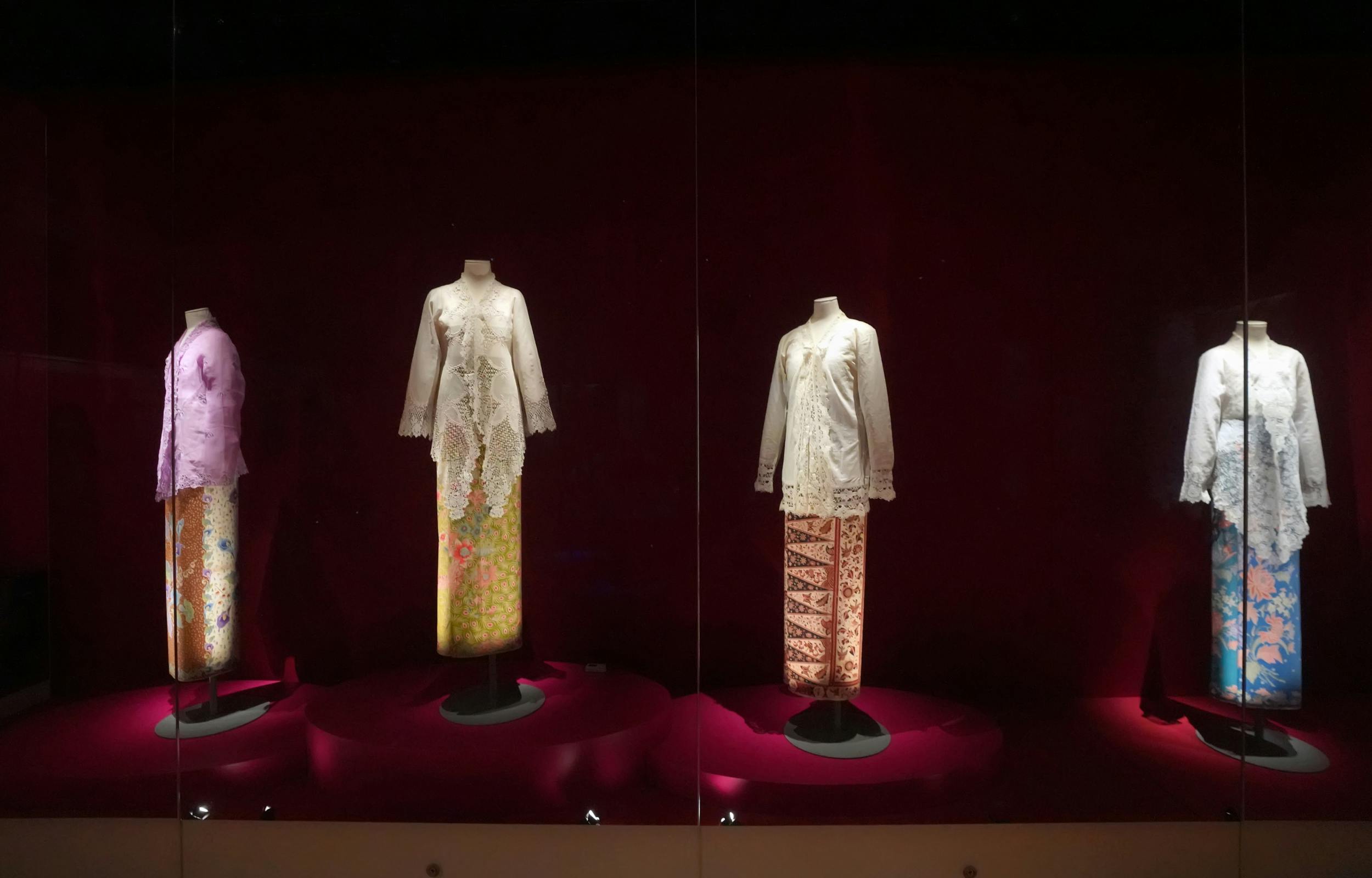 Two mannequins displaying traditional white lace blouses paired with patterned skirts, set against a red backdrop.