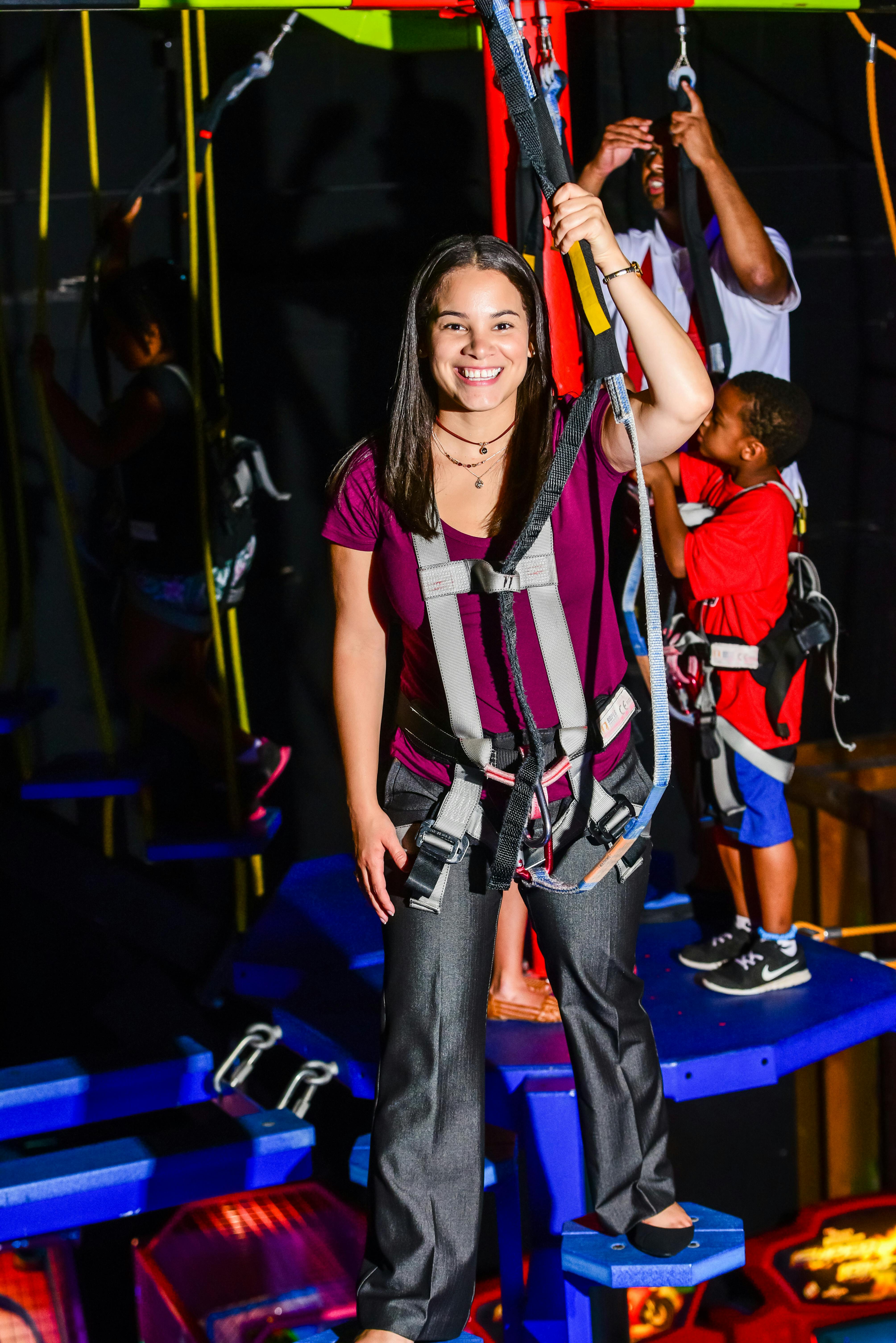 セーフティハーネスを着た女性が微笑み、他の人たちがロープコースを登っているのを背景にしています。