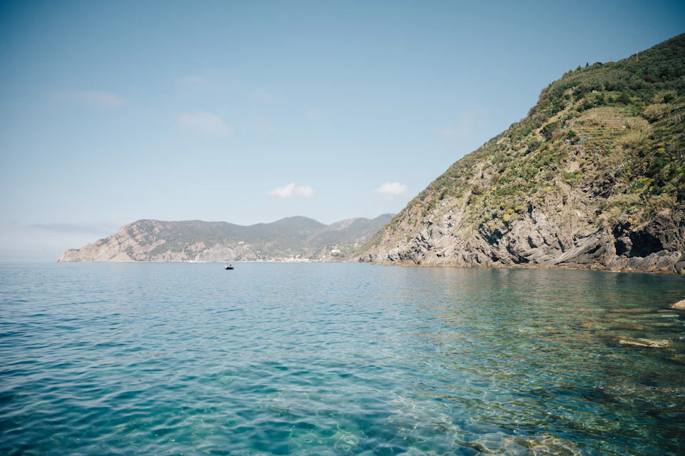 Clear blue sea with rocky shoreline on the right, distant mountains under a blue sky, and a small boat on the horizon.