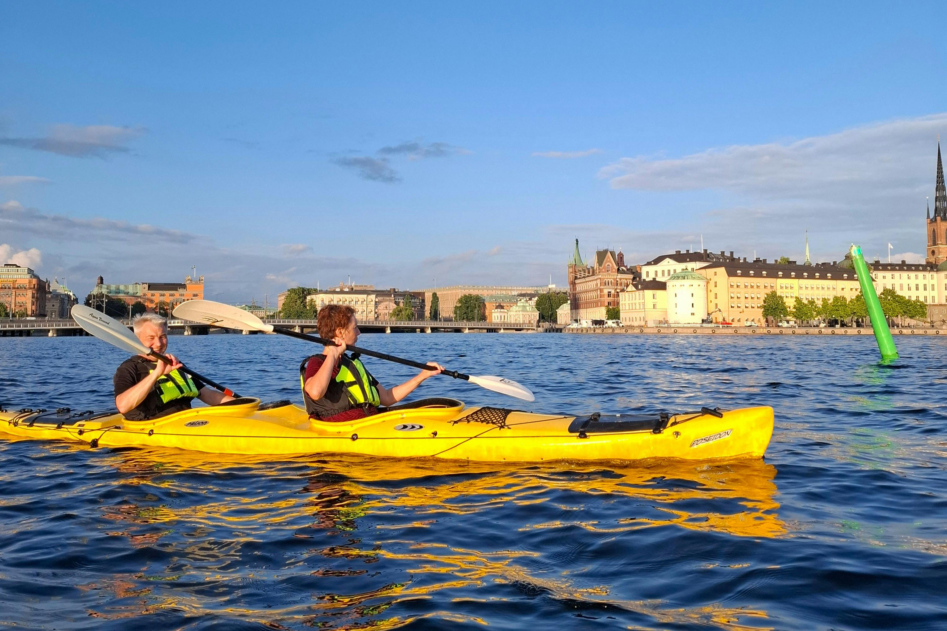 Deux personnes dans des kayaks jaunes pagaient sur un plan d'eau calme, avec des bâtiments urbains et un ciel bleu en arrière-plan.