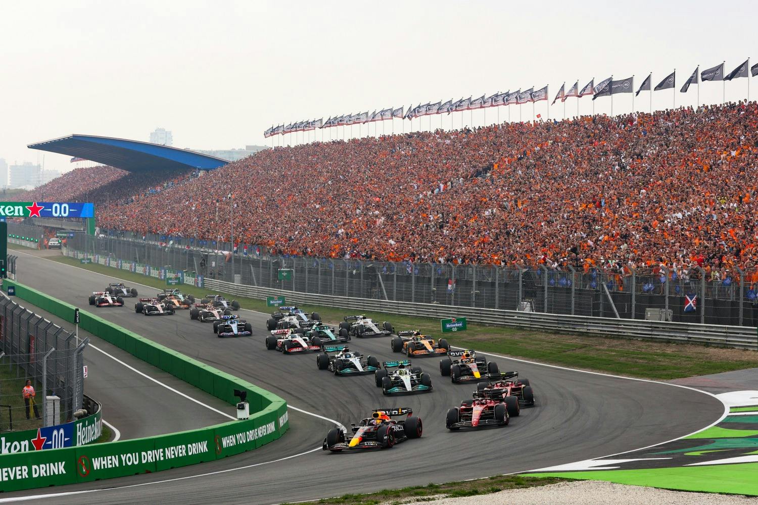 Racerbiler, der kører for hurtigt på en bane foran en fyldt tribune, med en Heineken-reklame og flag over publikum.
