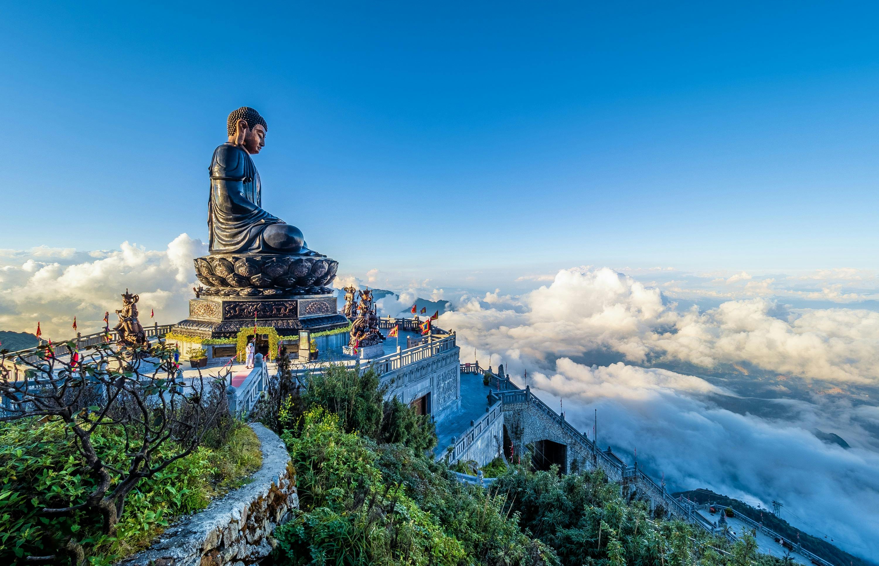 A large Buddha statue on a mountain terrace with surrounding greenery and clouds below, under a clear blue sky.