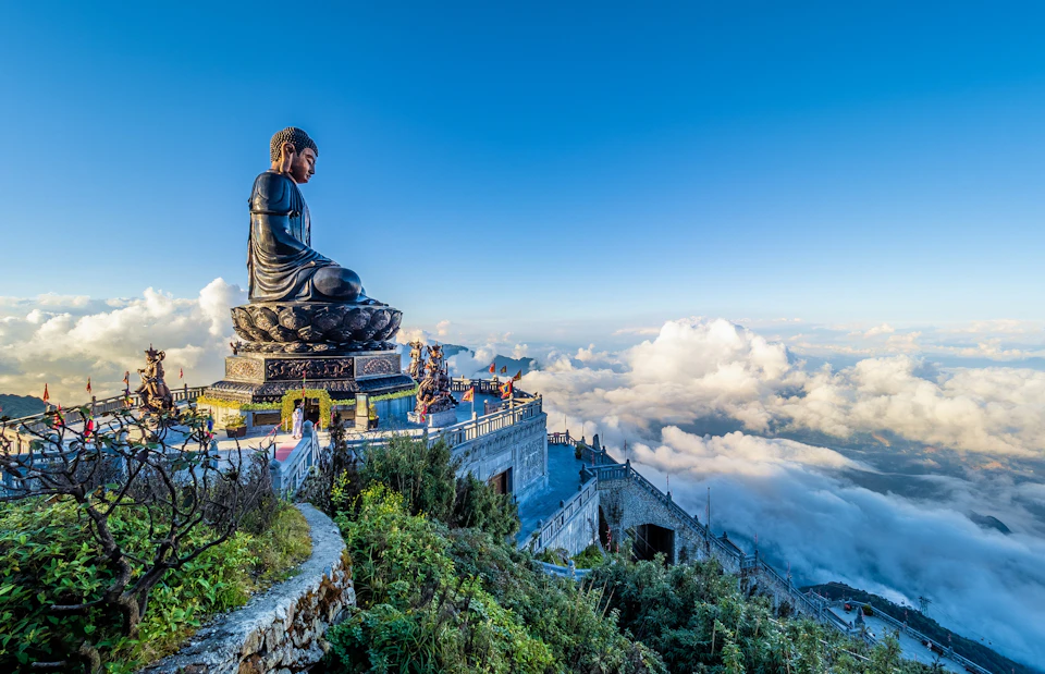 A large Buddha statue on a mountain, surrounded by clouds and greenery, with a scenic view and a clear blue sky.