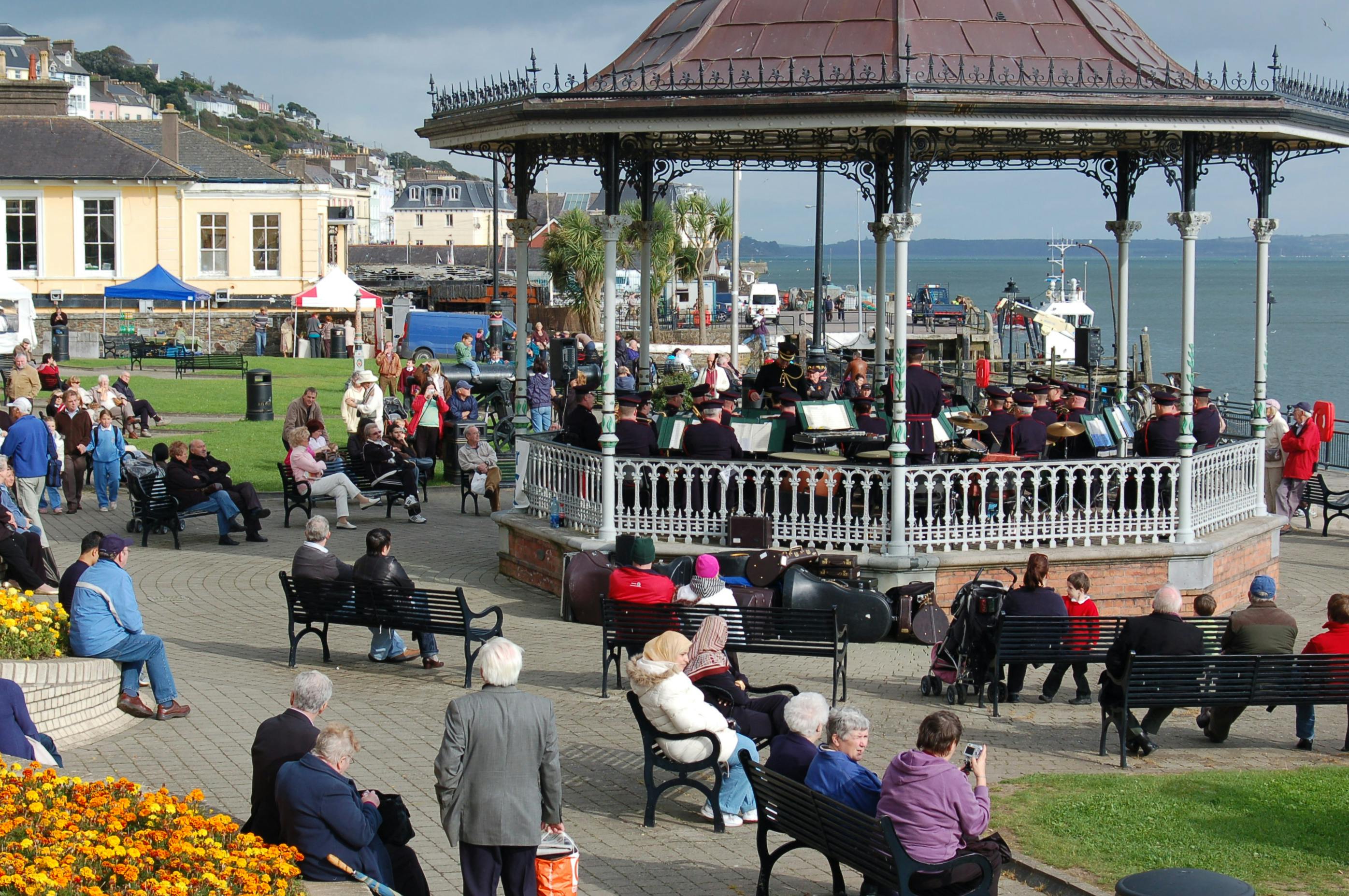People gathered around and sitting on benches near a gazebo with a band performing, set along a coastal town.