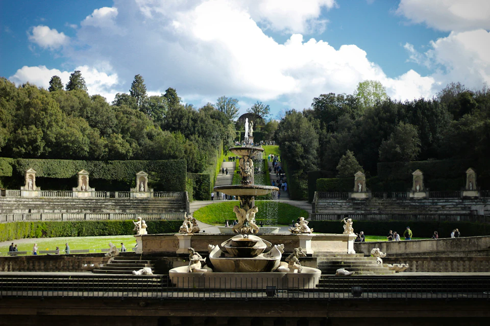 Fountain with statues and cascading water in an ornate, landscaped garden with trimmed hedges and trees under a partly cloudy sky.