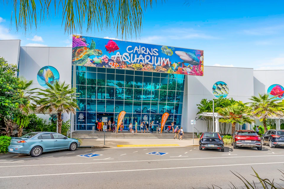 The entrance to Cairns Aquarium, featuring a colorful sign, glass facade, palm trees, parked cars, and visitors.