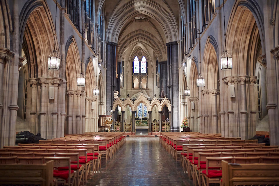 An empty church aisle with wooden pews and red cushions, stone arches, and an altar in the background.