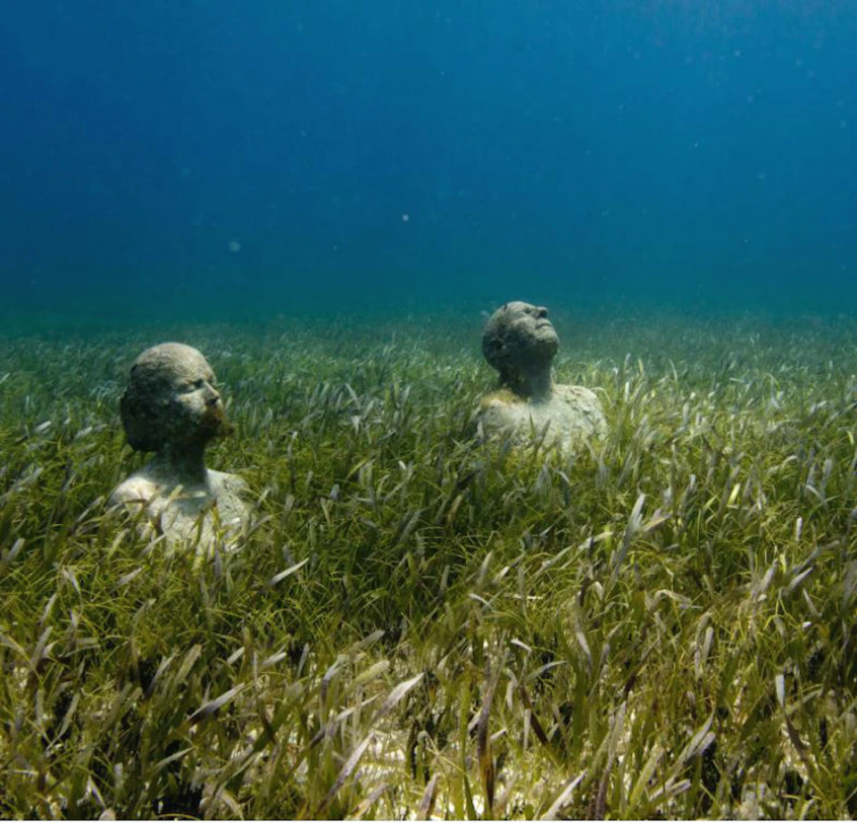 Two submerged stone busts of human heads and shoulders in an underwater landscape with seagrass and a blue water background.