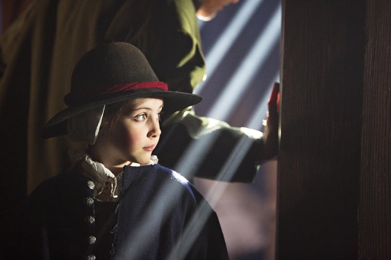 Dítě v historickém oděvu a klobouku se dívá do strany s paprsky světla osvětlujícími jeho tvář a částečně viditelnou osobou vedle nich.