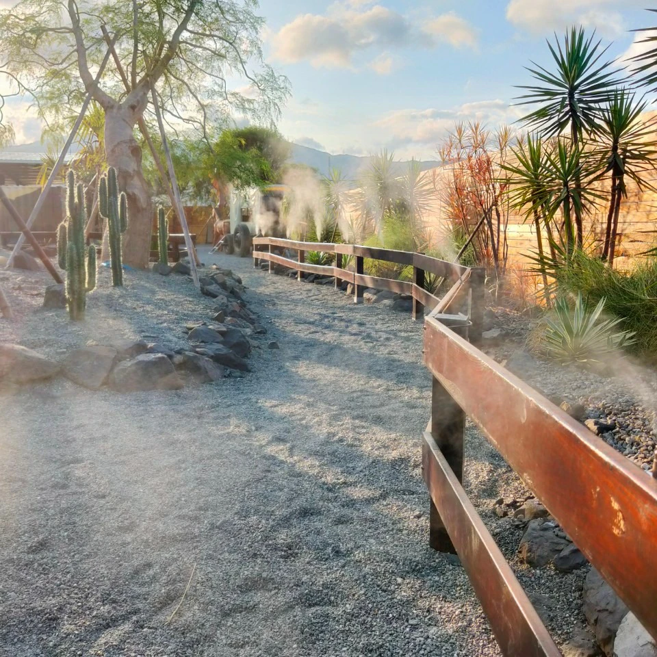 A gravel pathway with wooden railings bordered by dry landscape plants, including cacti, under a bright sky with scattered clouds.