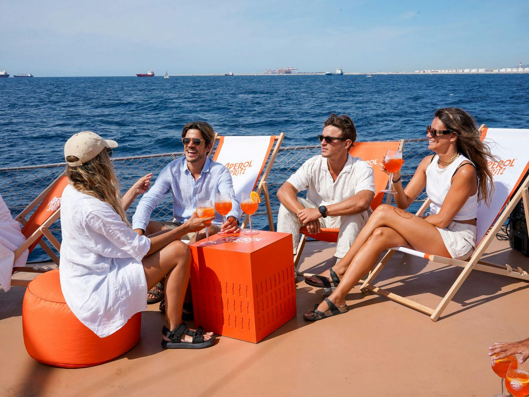 Quatre personnes se détendent sur un bateau, assises sur des chaises longues orange et blanches, en dégustant des boissons avec la mer et les bateaux en arrière-plan.