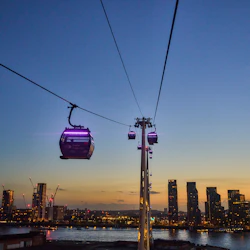 Ett linbanesystem över en flod i skymningen, med en stadssilhuett och upplysta byggnader i bakgrunden.