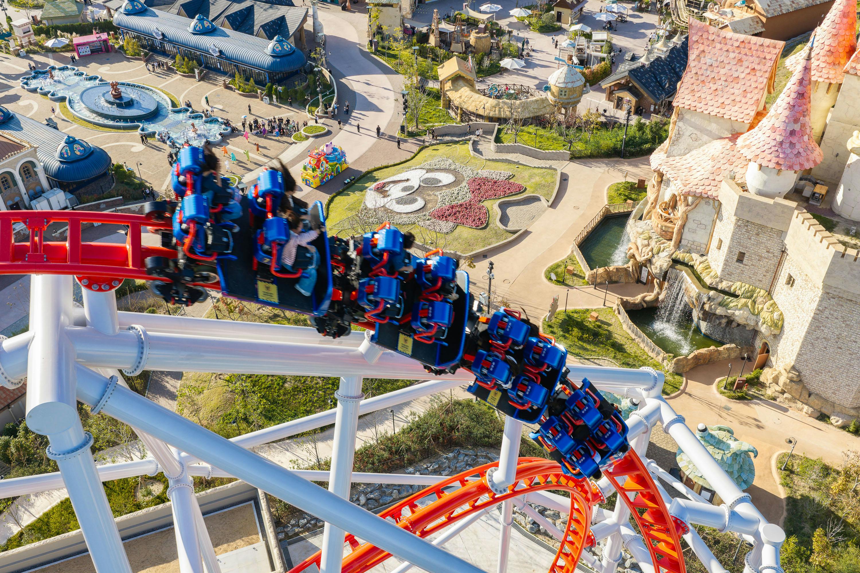 A rollercoaster with red rails and blue cars twists above an amusement park with themed buildings and a flower display.