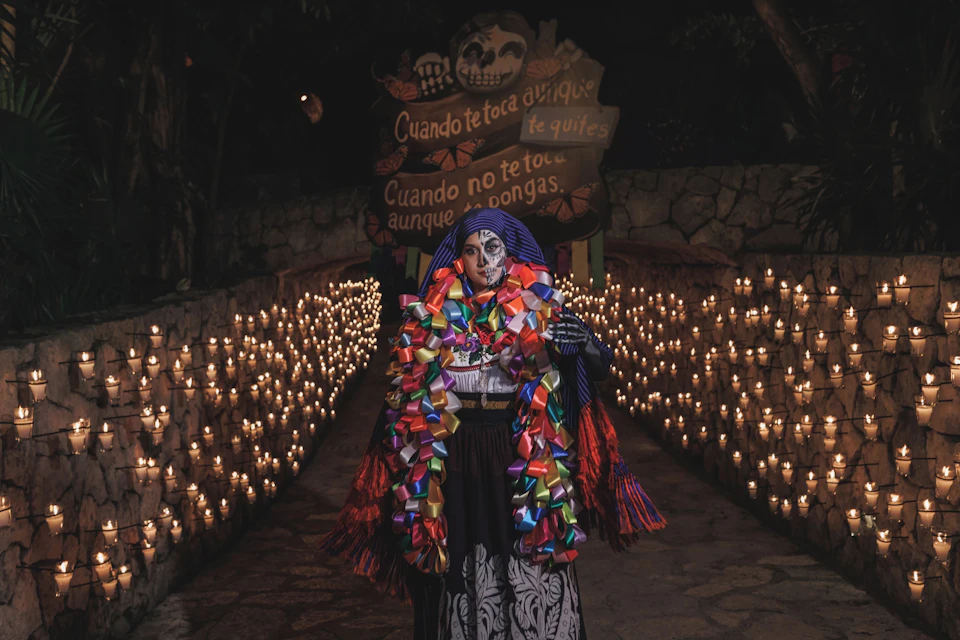 A person with face paint and colorful ribbons stands in a candle-lit pathway with a decorated sign in the background.