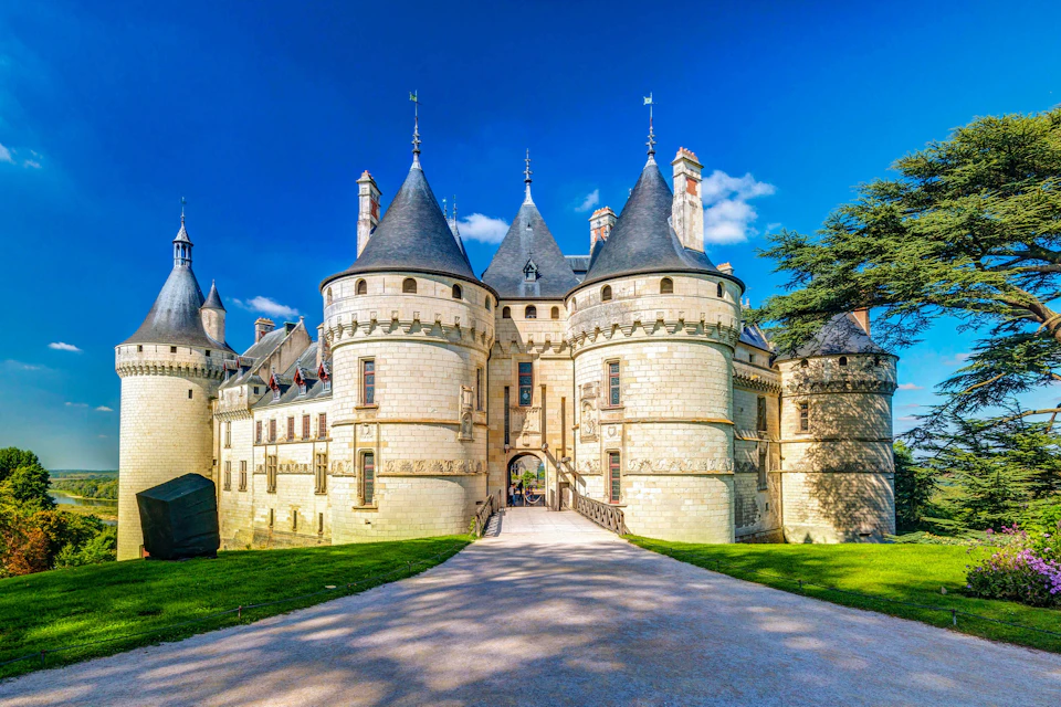 Stone castle with conical towers, a central archway, and a paved pathway leading to the entrance, under a clear blue sky.