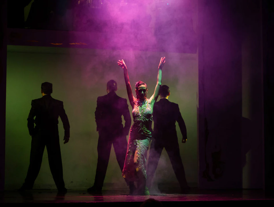A woman in a sequined dress dances with three men in suits against a smoky, purple-lit background.