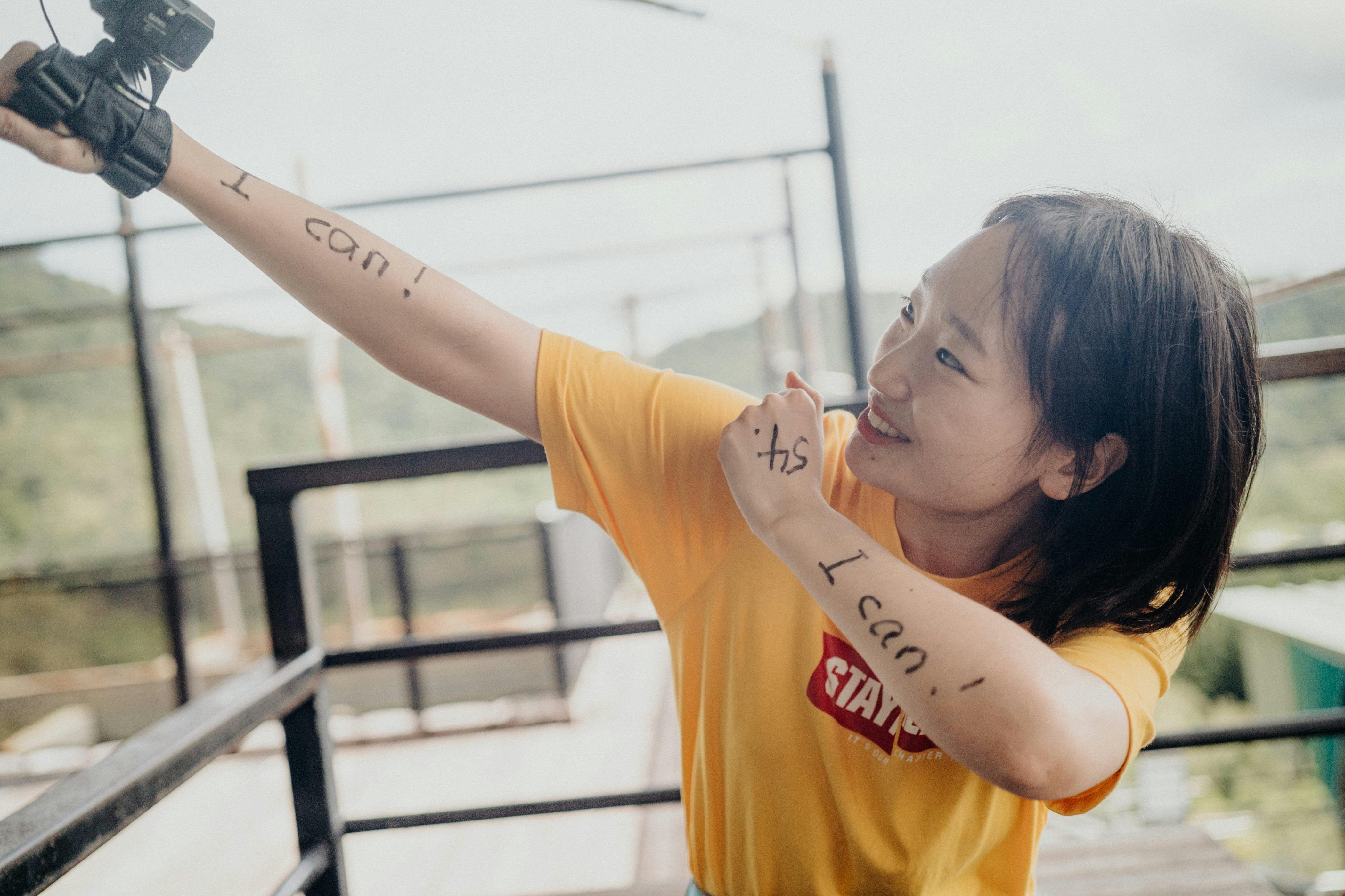 A person in a yellow shirt with "I can!" written on their arms holds a camera, smiling energetically outdoors.