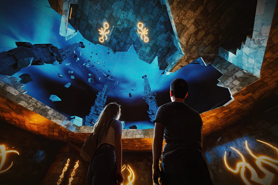 Two people stand in a room with a cave-like ceiling displaying a blue, intricate design with glowing symbols and stone structures.