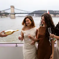 Duas mulheres a sorrir, segurando copos de champanhe num barco, com uma ponte e um rio ao fundo.