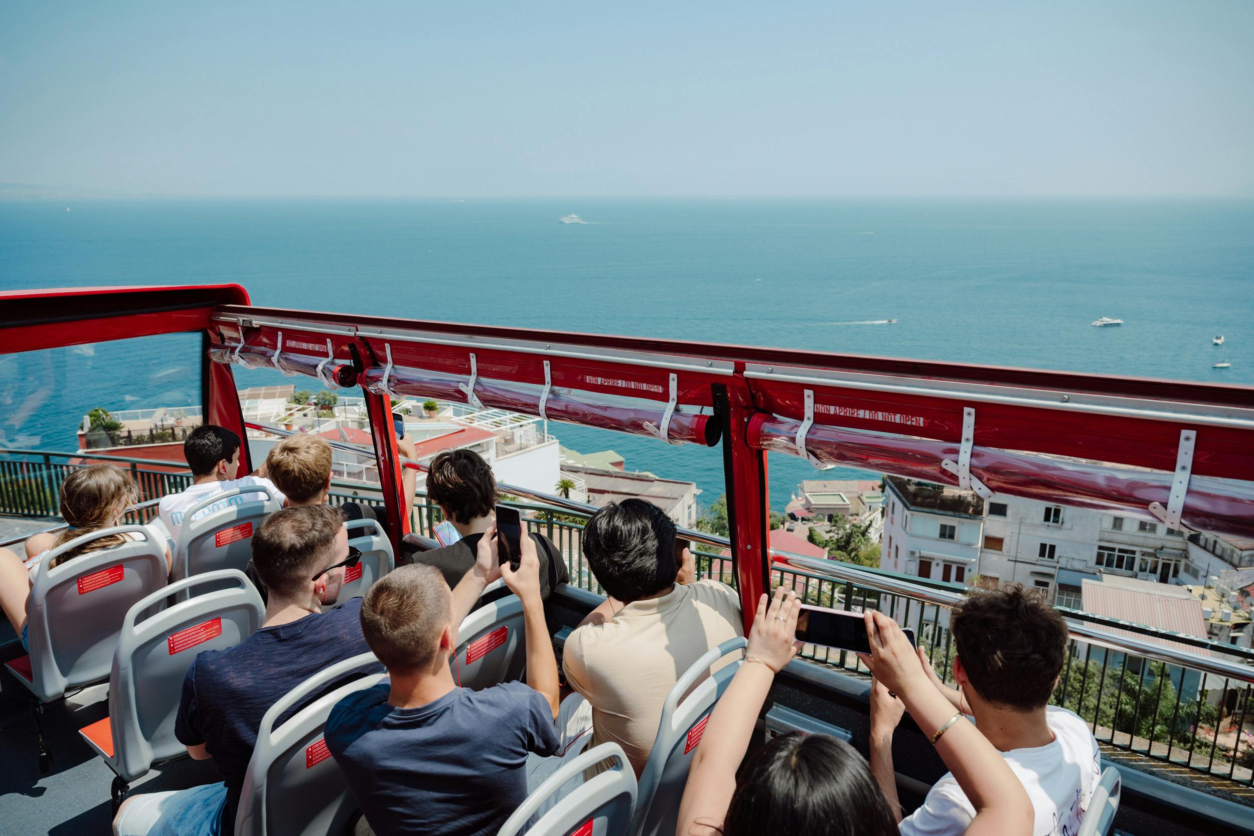 Persone sedute su un autobus scoperto che fotografano una vista panoramica della costa con edifici e l'oceano visibili.