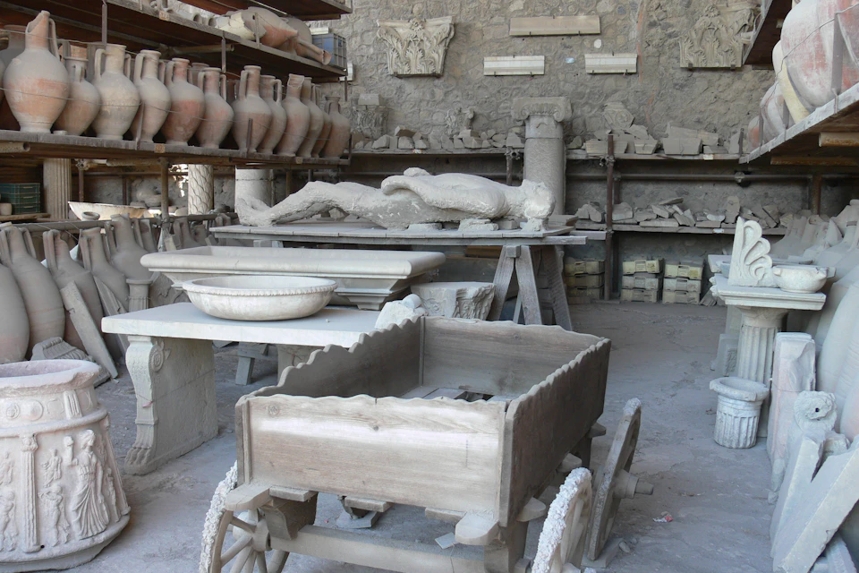 A room with ancient artifacts, including pottery on shelves, a wooden cart, and plaster casts of human figures on tables.