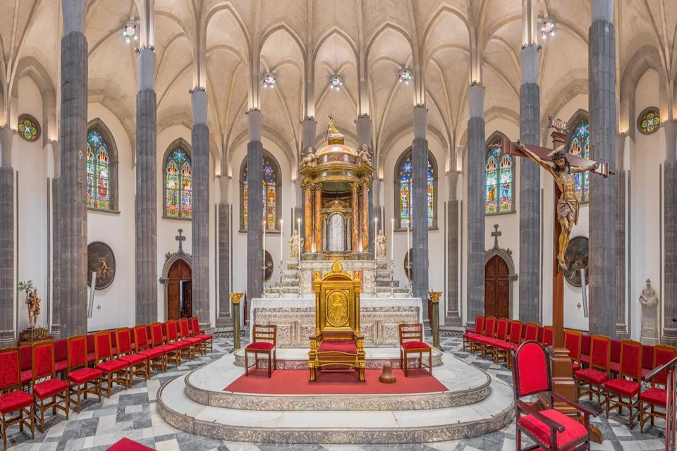 An ornate church interior with a central altar, stained glass windows, and red-seated chairs arranged in rows.